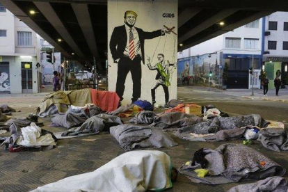 Un grupo de sin techo duerme bajo un puente de Sao Paulo ante una pintada de Bolsonaro como marioneta de Trump.