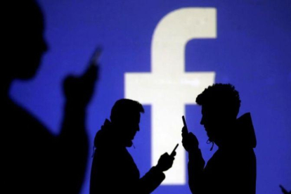 Usuarios consultan su móvil delante de una pantalla en la que se proyecta el logo de Facebook.