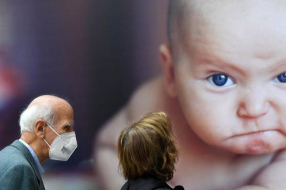Dos personas con mascarilla pasan por delante de un cartel con la imagen de un bebé.  DAL ZENNARO