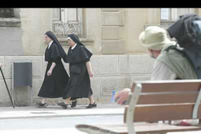 Una pareja de monjas camina por la calle frente a un peregrino que descansa sentado en un banco.