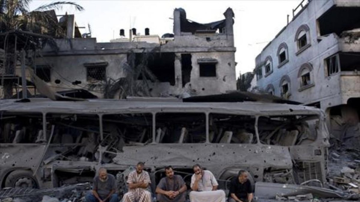 Cinco palestinos de Gaza sentados frente a un autobús y un edificio destruidos, en el 2014, por ataques aéreos israelís.