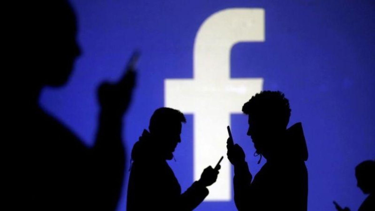 Usuarios consultan su móvil delante de una pantalla en la que se proyecta el logo de Facebook