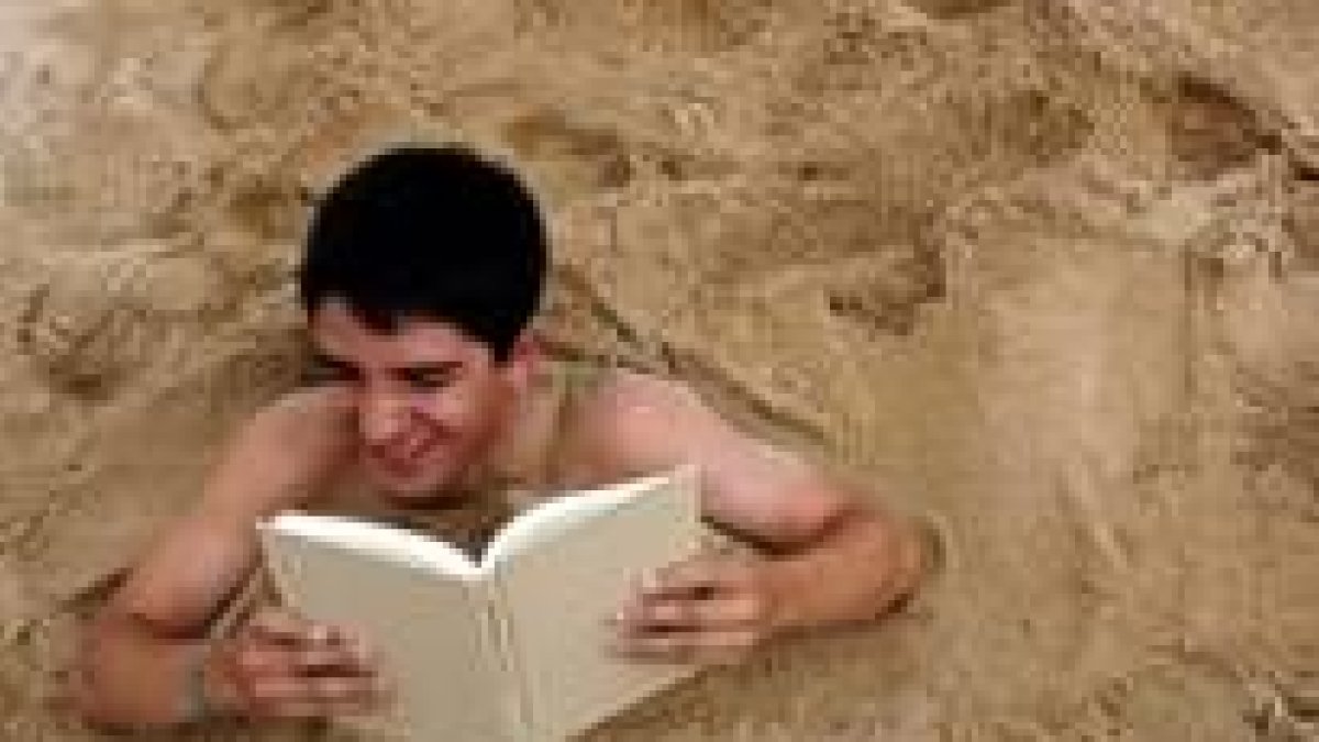 Un joven lee un libro mientras permanece semienterrado en la arena de una playa del Atlántico