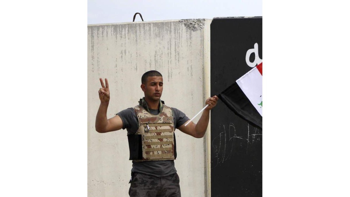 Un miembro del ejército iraquí con la bandera nacional; a la derecha, un hombre sostiene un cartel.
