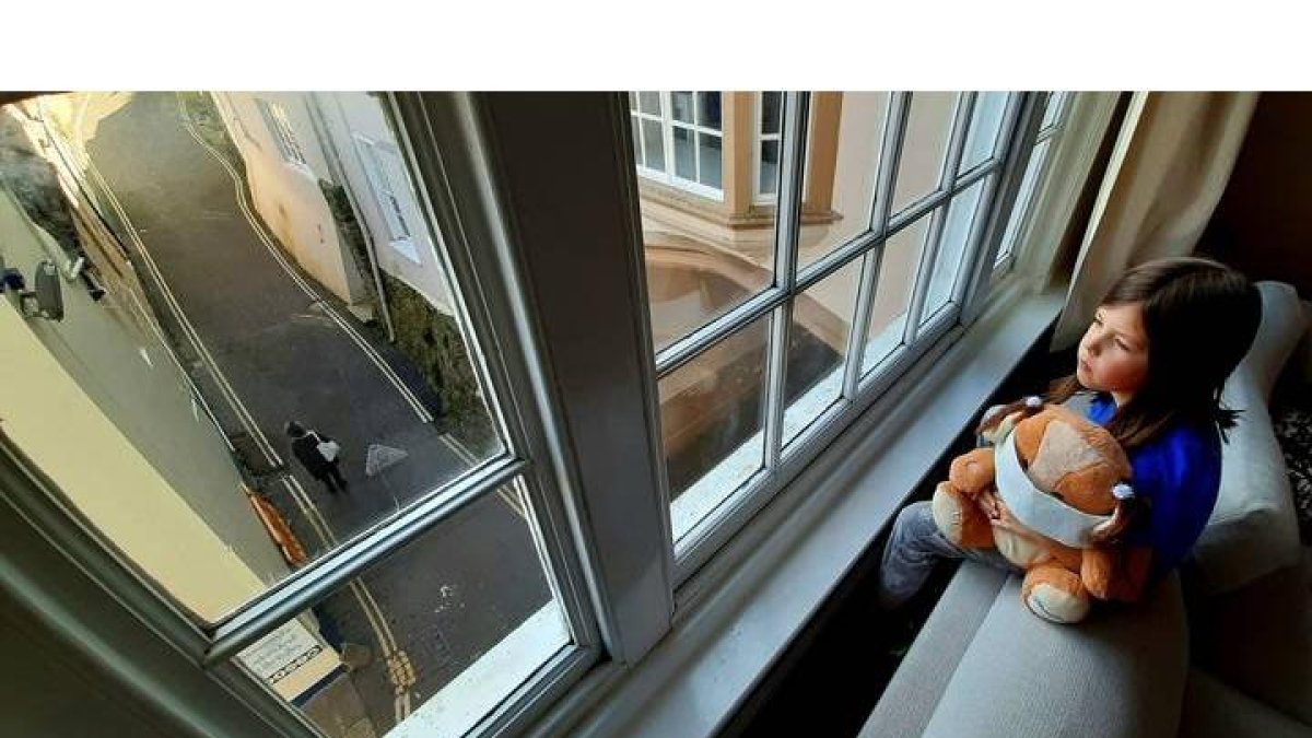 Una niña abraza a su oso de pelucho, ataviado con una máscara sanitaria, mientras miran por la ventana de su casa. ROMAN ALONSO