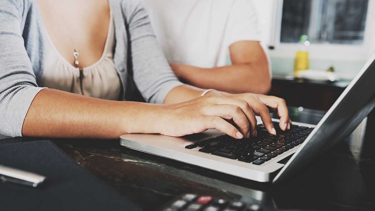Hands of young woman working on laptop at home with husband sitting next to her