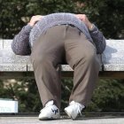 Una persona aprovecha el sol para dormir una siesta en un banco de una calle.