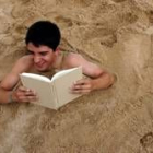 Un joven lee un libro mientras permanece semienterrado en la arena de una playa del Atlántico