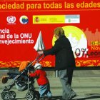 Una señora pasea a un niño pequeño delante de un cartel que anuncia un encuentro en León sobre envej
