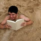 Un joven lee un libro mientras permanece semienterrado en la arena de una playa del Atléntico