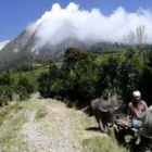 Un hombre conduce un carrito tirado por un búfalo mientras el humo del volcán cubre el distrito.