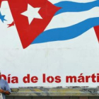 Un hombre lee un periódico junto a un cartel con una bandera cubana.