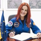 La astronauta Sara García‑Alonso firma el libro de visitas del SatCen vestida con su traje de la ESA, flanqueada por las banderas de España y la Unión Europea.