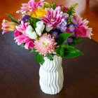 Un ramo vibrante con lirios, rosas y crisantemos en jarrón blanco que transforma cualquier mesa en un punto focal lleno de color y frescura.