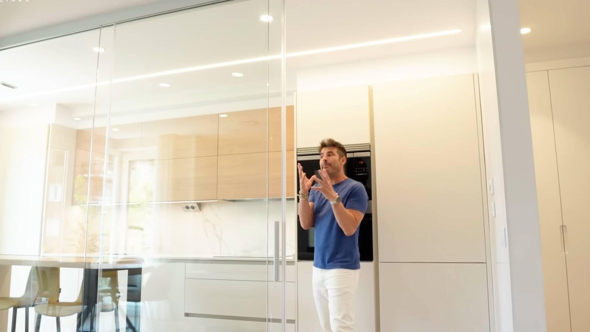 Vista frontal de la cocina reformada por el estudio de Daniel Colino, con materiales como madera y blanco perla y una división en cristal que conecta con el salón.