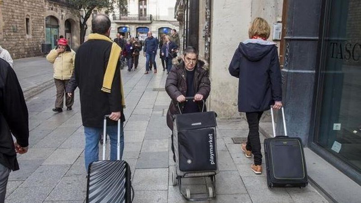 Una mujer pasa con su carrito de la compra entre dos turistas con sus maletas.