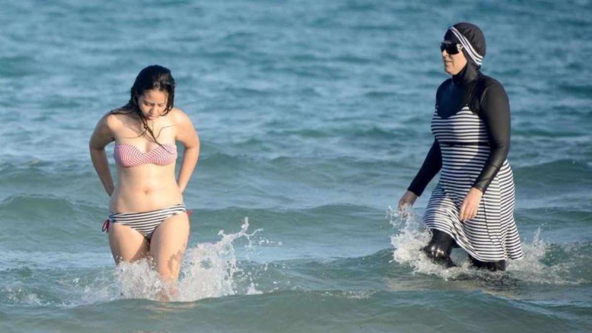 Una mujer con biquini y otra con 'burkini' en la playa tunecina de Ghar El Melh.