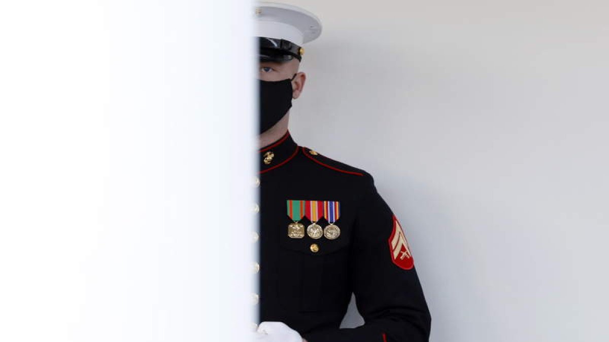 Un marine custodia la puerta principal de la Casa Blanca horas antes de la llegada del nuevo presidente, Joe Biden. Miles de banderas jalonan la avenida que lleva al Capitolio la víspera de la toma de psosesión de Biden. Jacob Anthony Chansley durante el asalto a la Cámara de Representantes. En la otra página, tropas con trajes de época marchan en el ensayo del traspaso de poderes