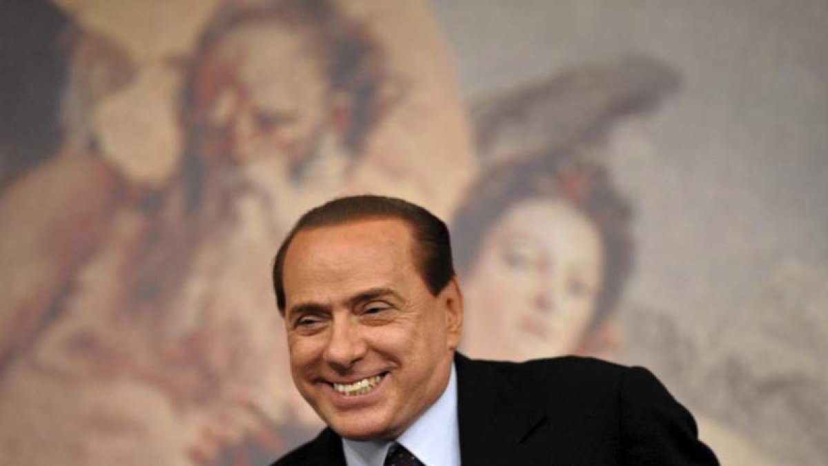 Silvio Berlusconi sonríe mientras toma asiento en una rueda de prensa.