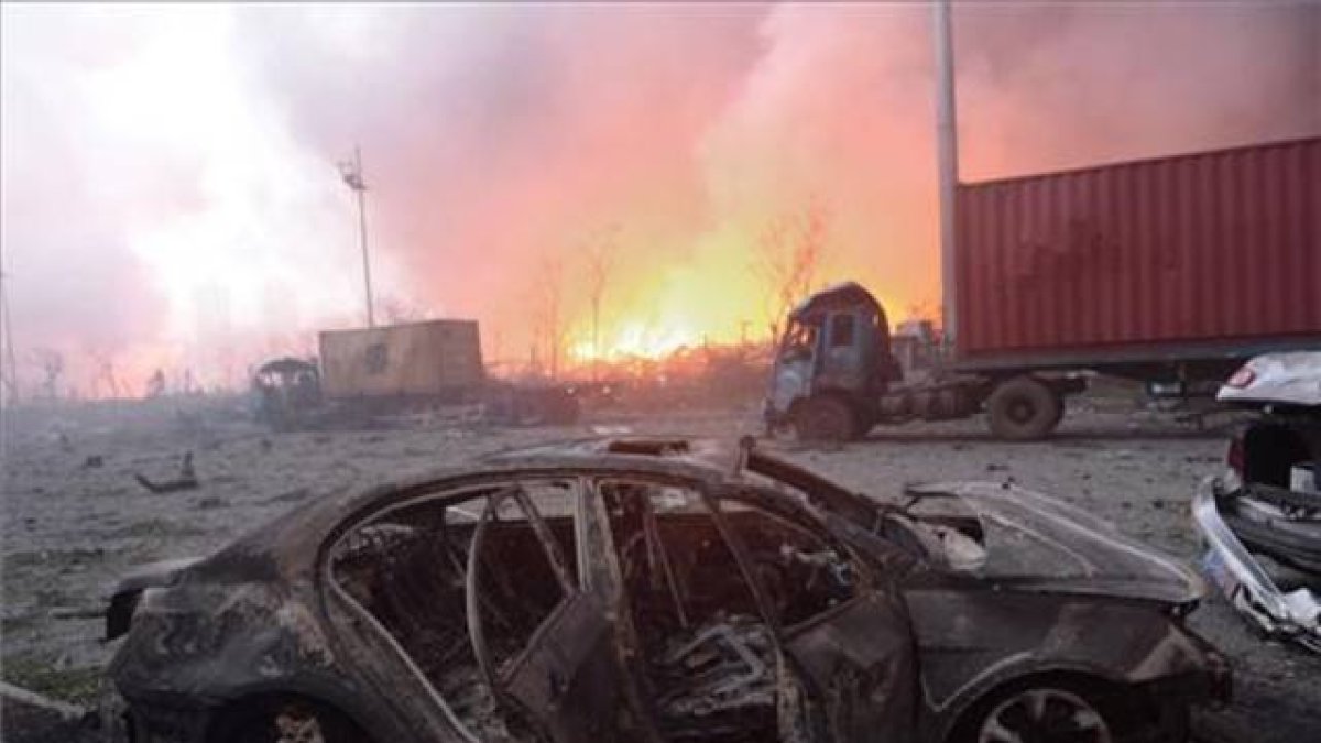 Vista de un coche calcinado en la seria de explosiones de unos almacenes de Tianjin, en China. En segundo plano, el fuego sigue ardiendo.