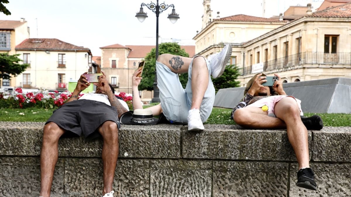 Tres jóvenes consultan el móvil tumbados en la calle este domingo en el centro de León.