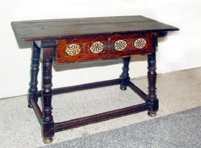 Mesa de forma rectangular con cuatro montantes tipo balaustres torneados, alternando figuras cilíndricas con jarrón invertido, unidos por cuatro travesaños lisos. Peinazos muy gruesos con guías interiores. Obra entregada en calidad de depósito por la Comisaría General del Patrimonio Artístico Nacional en 1939.