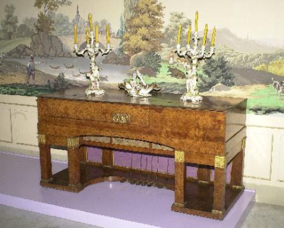 El austríaco Ignace Joseph Pleyel (1757-1831), compositor y pianista afincado en París, se asoció en 1805 con Lemme, constructor alemán de pianos, para fundar una empresa que fabricara estos instrumentos.Mueble de estilo imperio chapado en madera de raíz y sustentado por cuatro parejas de patas de sección cuadrangular en el frente y en la parte posterior, con aplicaciones de bronce dorado a modo de capiteles de columnas que se completan con los roleos vegetales y los motivos antropomorfos de la parte central de la tapa abatible. Obra entregada en calidad de depósito por la Comisaría General del Patrimonio Artístico Nacional el 28 de noviembre de 1939.