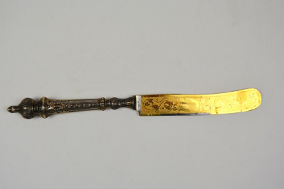 Cuchillo compuesto de mango en color plateado y hoja dorada. El mango presenta un talón con un pequeño pomo gallonado con una protuberancia en el remate. Obra entregada en calidad de depósito por el Ministerio de Educación y Ciencia, el 24 de febrero de 1977.